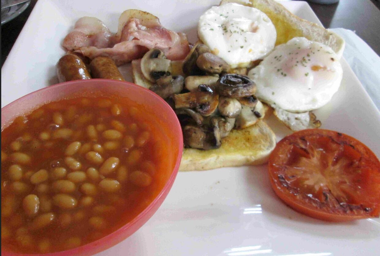 Un plato con frijoles, salchichas, tocino, huevos, champiñones, tostadas y una rodaja de tomate.