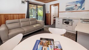 Una habitación moderna con un sofá color beige, una mesa de comedor blanca con revistas, una zona de cocina y vistas al exterior.