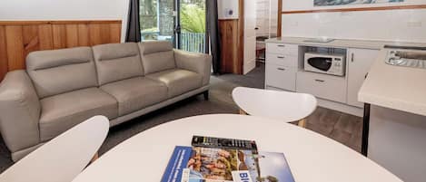 Una habitación moderna con un sofá color beige, una mesa de comedor blanca con revistas, una zona de cocina y vistas al exterior.