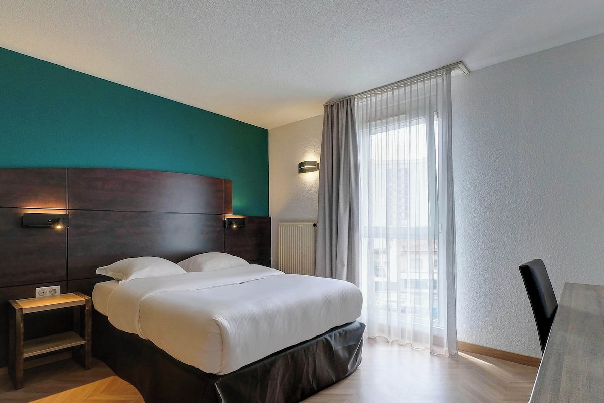 A hotel room with a large bed, a bedside table, a nightstand, a chair, and a window with curtains.