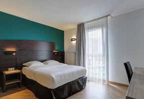 A hotel room with a large bed, a bedside table, a nightstand, a chair, and a window with curtains.