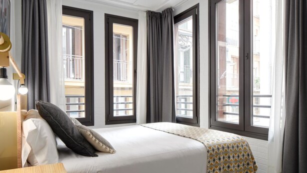 Habitació d'hotel amb llit, escriptori i grans finestres amb cortines.