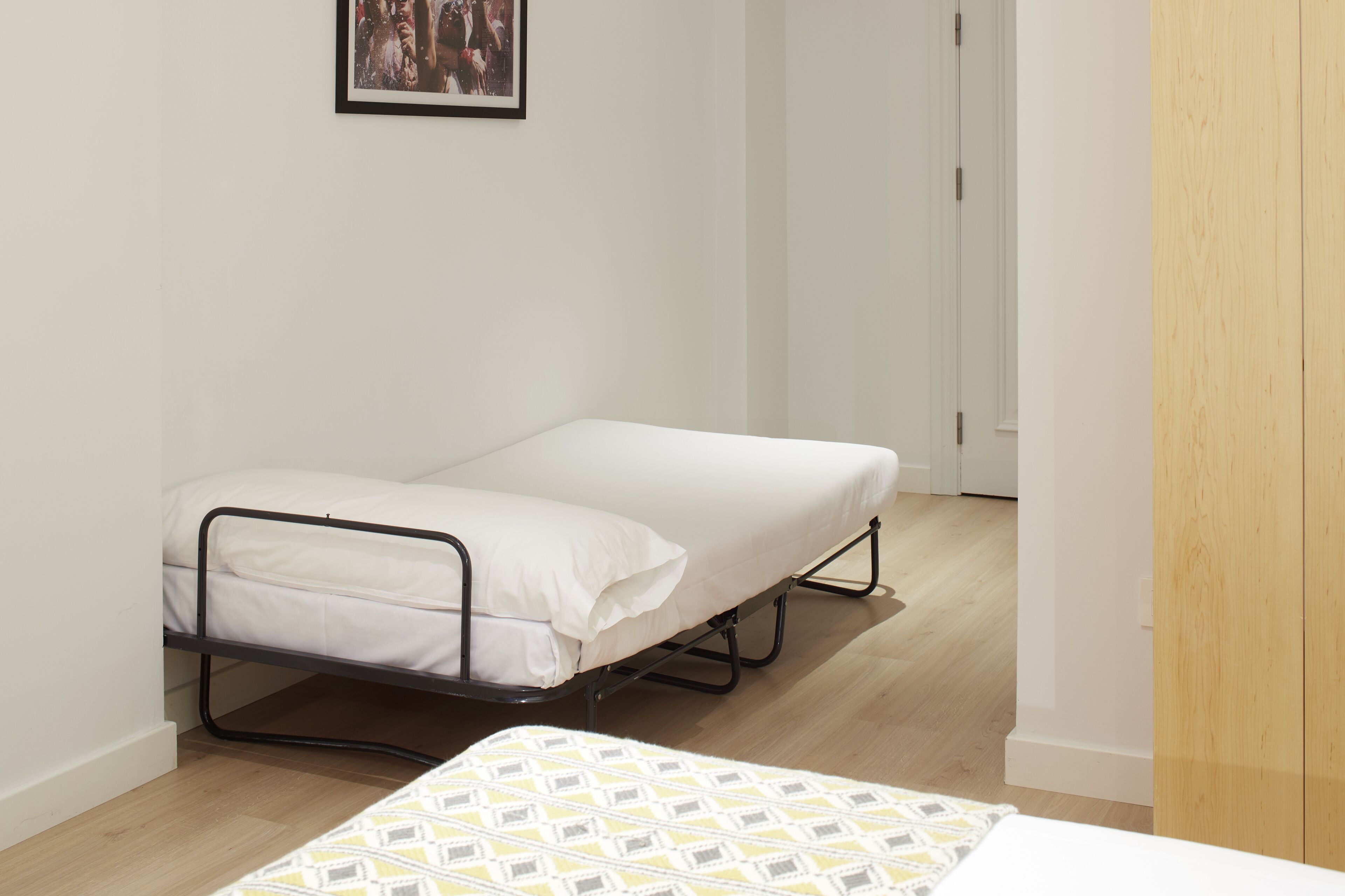 Une petite chambre avec un lit simple, un cadre de lit en métal, du linge de lit blanc et un plancher en bois.