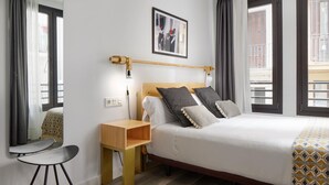 Habitació d'hotel amb llit, taulella de nit, cadira i finestra amb cortines.