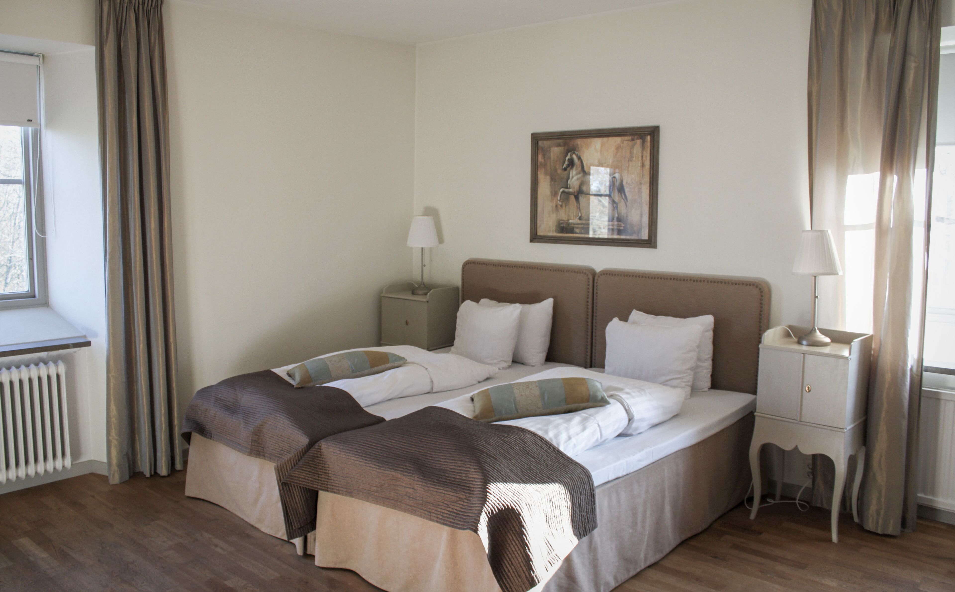 A hotel room with a large bed, two bedside tables, a radiator, and a window with curtains.