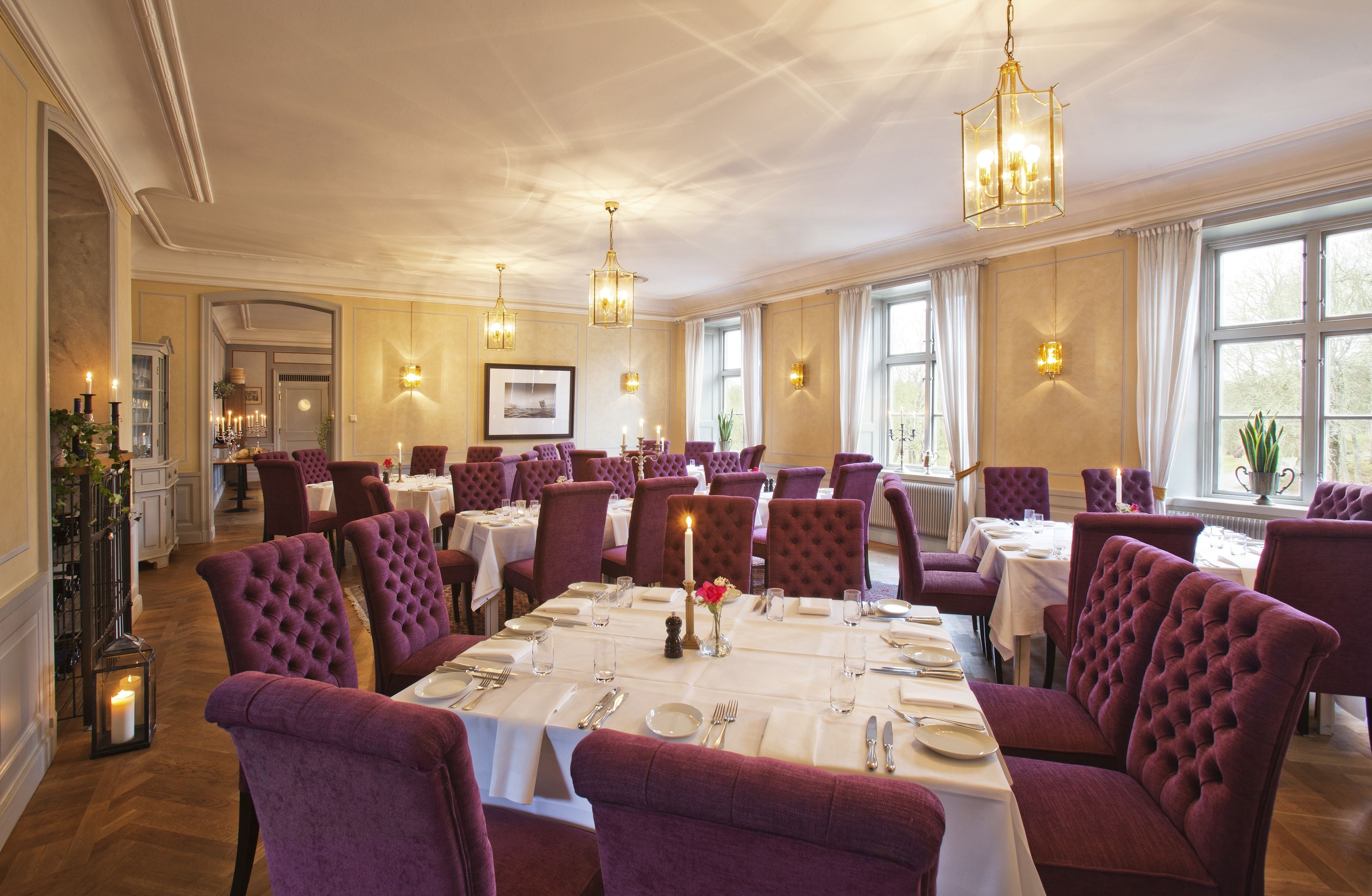 A formal dining room with multiple tables and chairs, elegant chandeliers, and large windows.