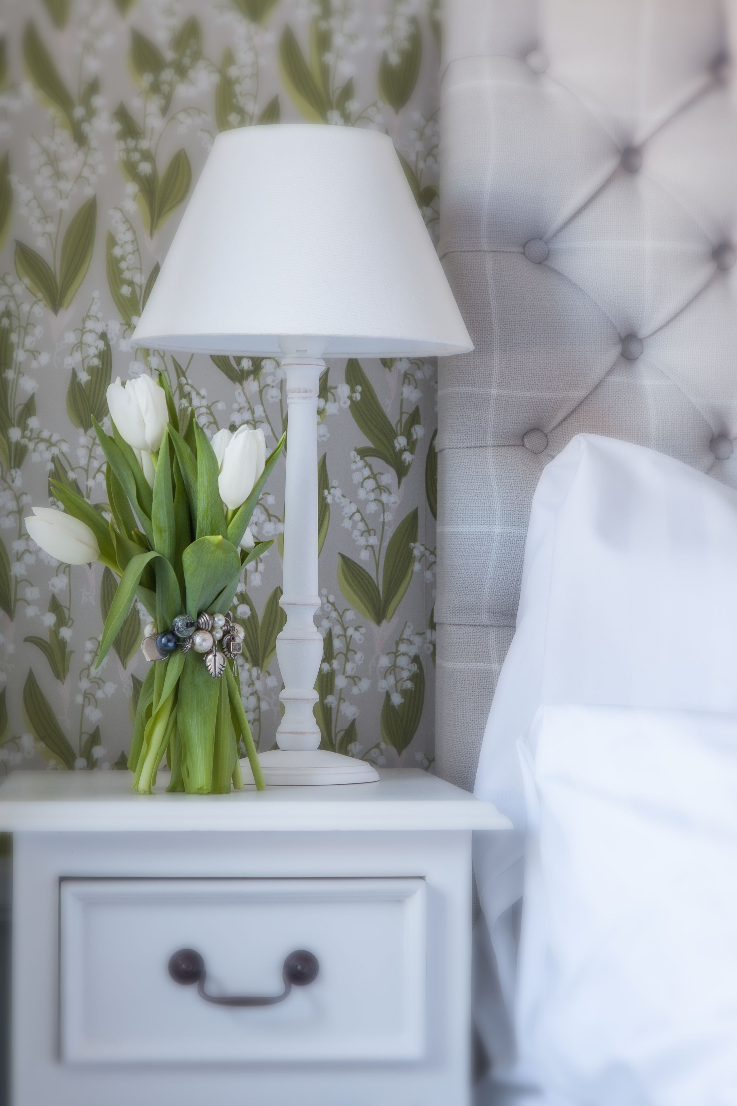 Una mesita de noche blanca con una lámpara y un ramo de flores blancas.