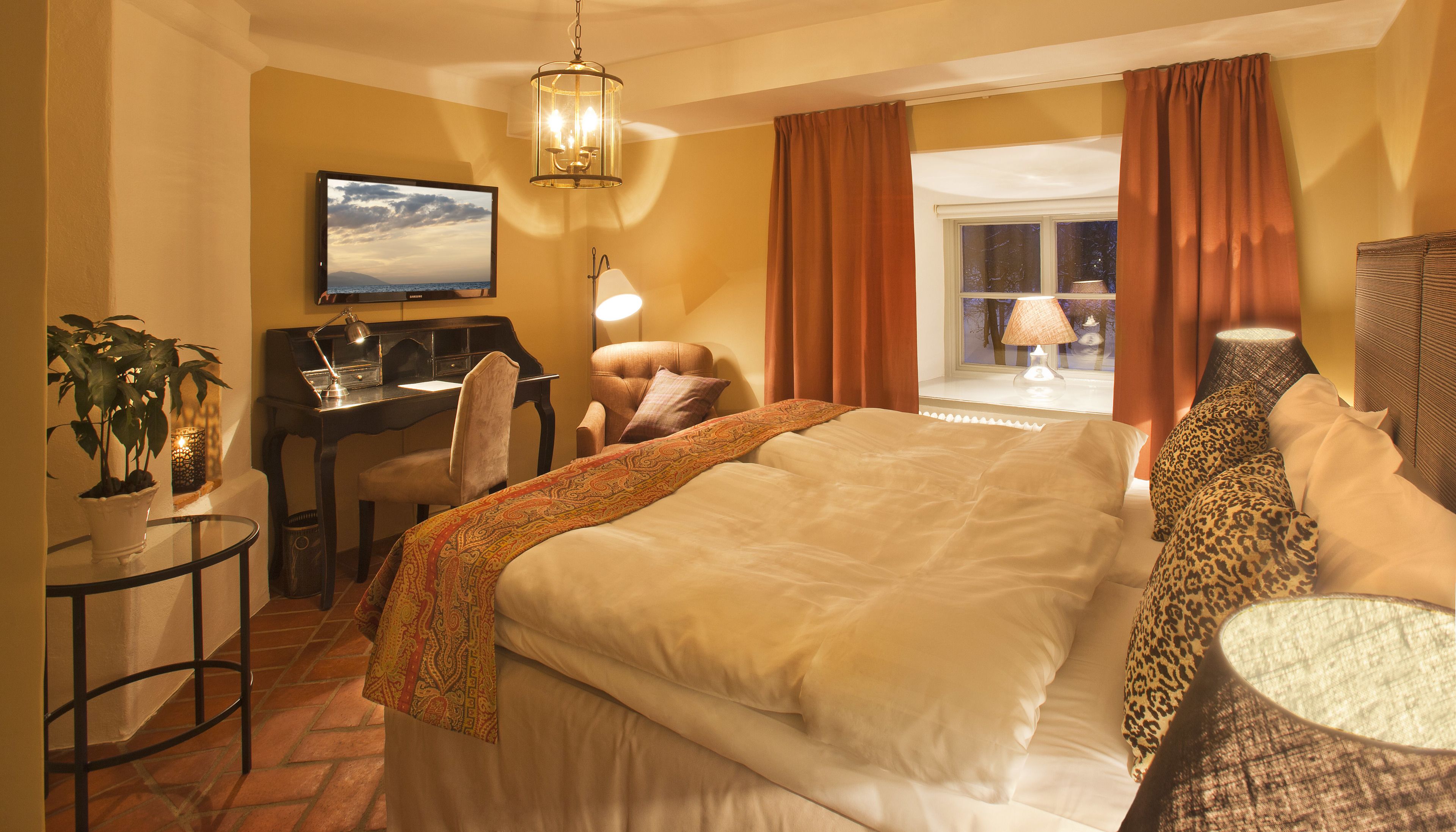 A hotel room with a large bed, a desk with a computer, a chair, a potted plant, and a view of the outdoors through a window.