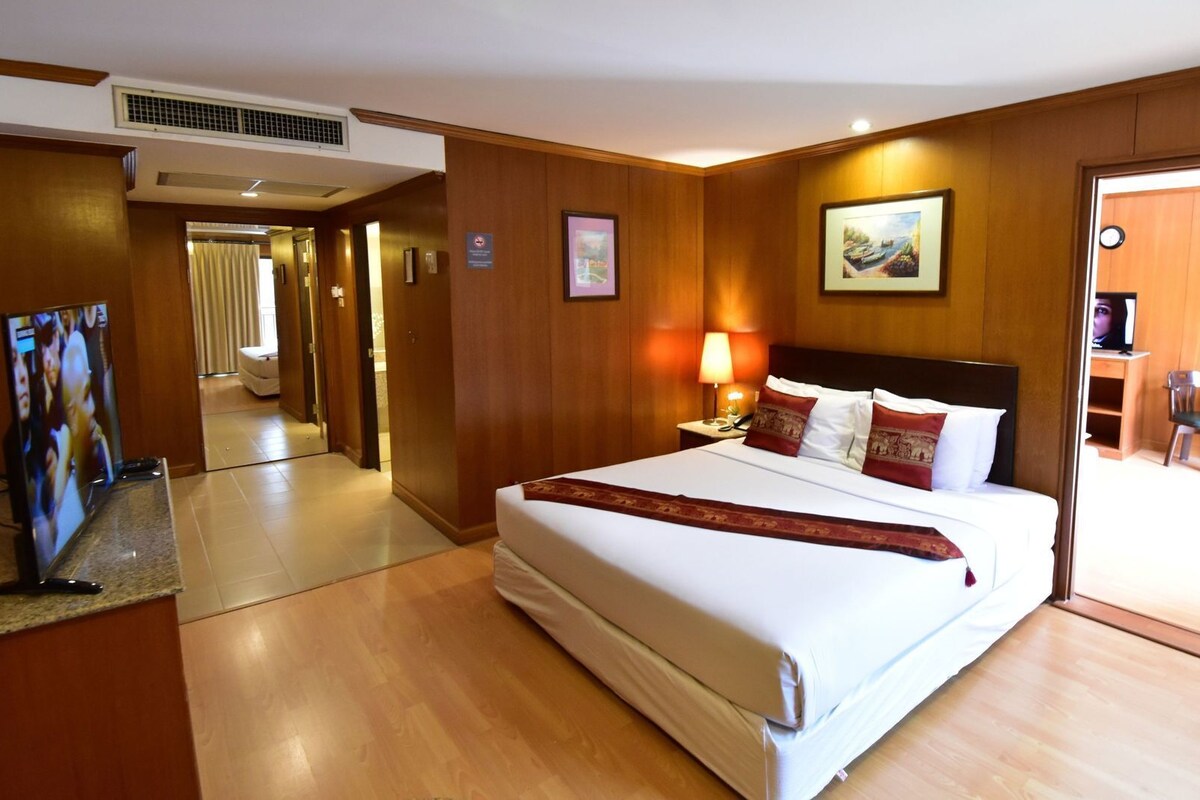 A hotel room with a large bed, a bedside table with a lamp, a TV, and a door leading to a dining area.