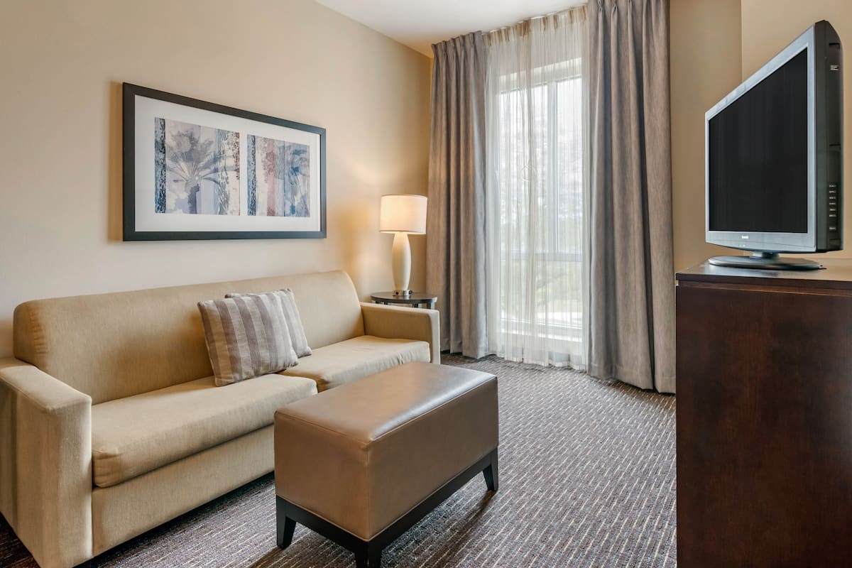 A hotel room with a beige sofa, a small ottoman, a flat-screen TV on a wooden stand, a framed abstract art piece on the wall, a side table with a lamp, and a large window with curtains.