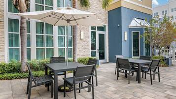 Un patio al aire libre con una mesa, sillas, un paraguas grande y un edificio con ventanas de piedra y vidrio.