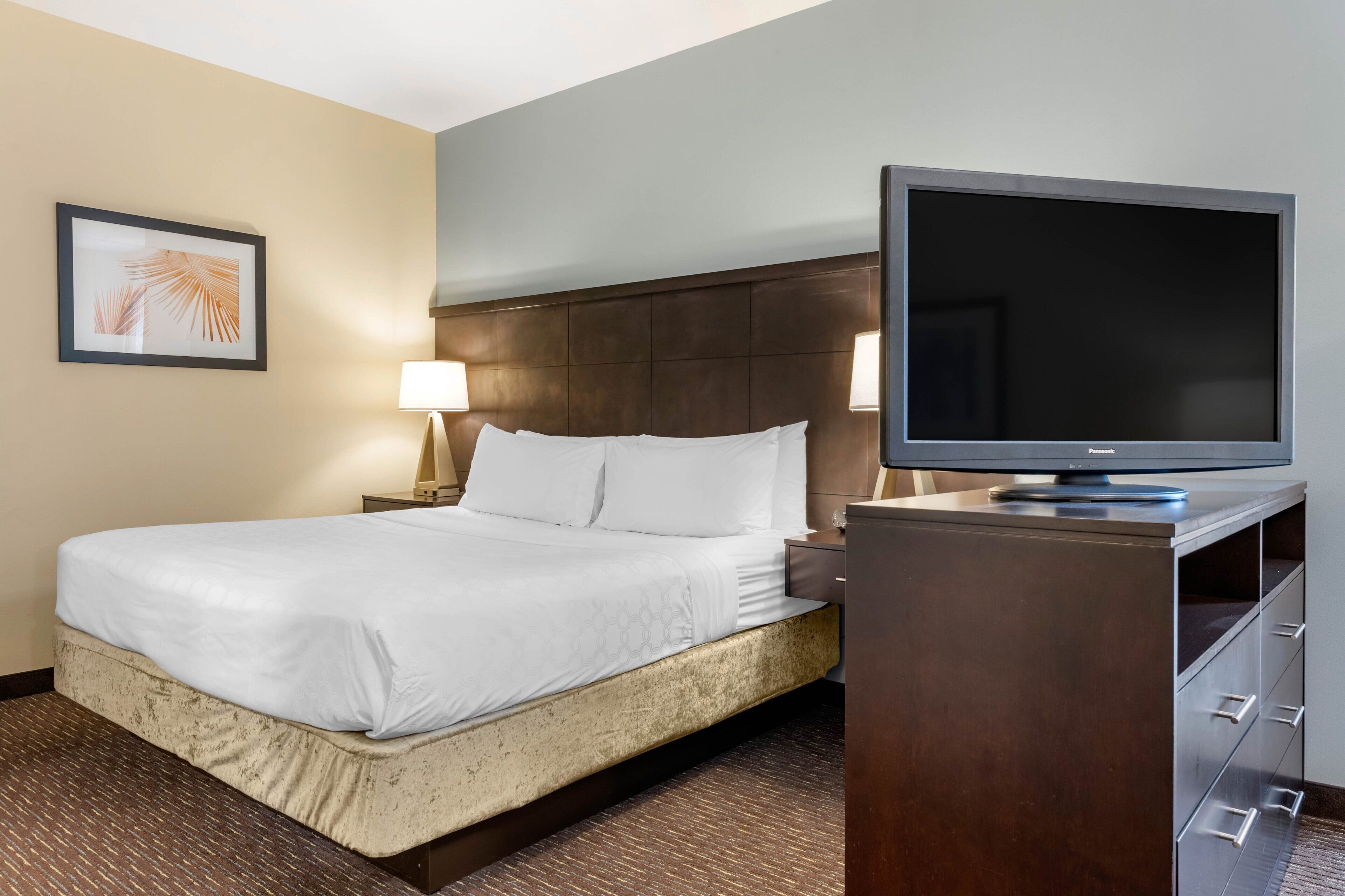 A hotel room with a bed, a television on a stand, a nightstand, a lamp, and a framed picture on the wall.