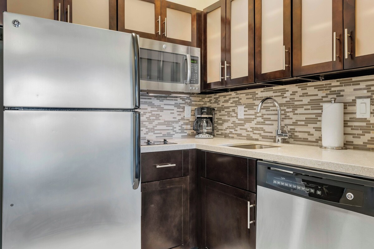 A kitchen with stainless steel appliances, dark wooden cabinets, a tiled backsplash, and a dishwasher.