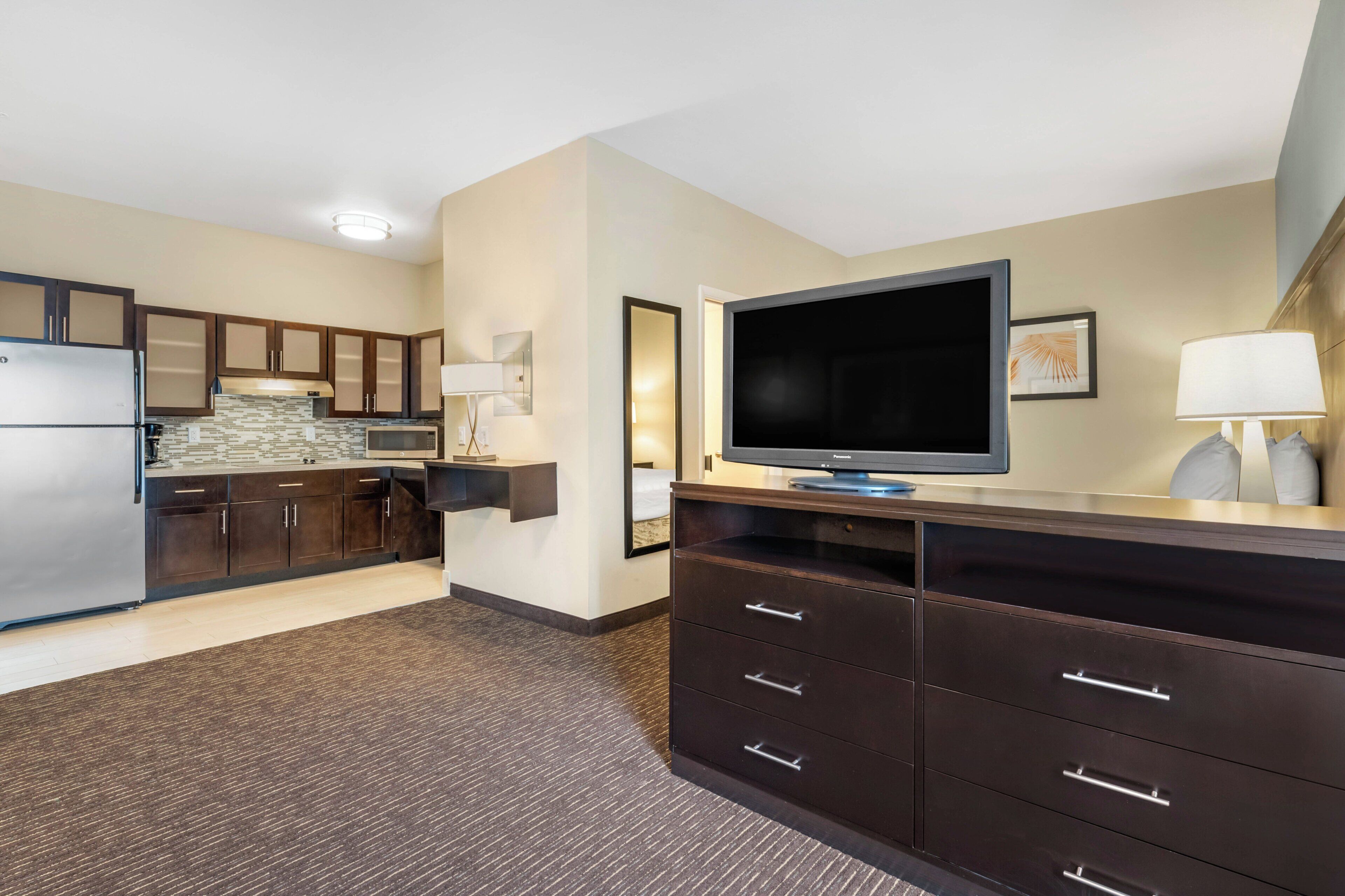 Quarto de hotel moderno com cozinha compacta, televisão de tela plana, espelho grande e cômoda de madeira escura.