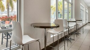 A modern interior with a long bar area, white chairs, and large windows offering a view of palm trees and a building outside. - Staybridge Suites St. Petersburg Downtown by IHG (St. Petersburg)