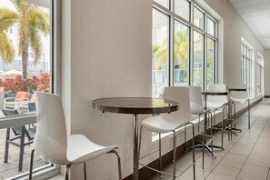 Un interior moderno con una barra alargada, sillas blancas y ventanas amplias que ofrecen vista a palmeras y un edificio exterior.