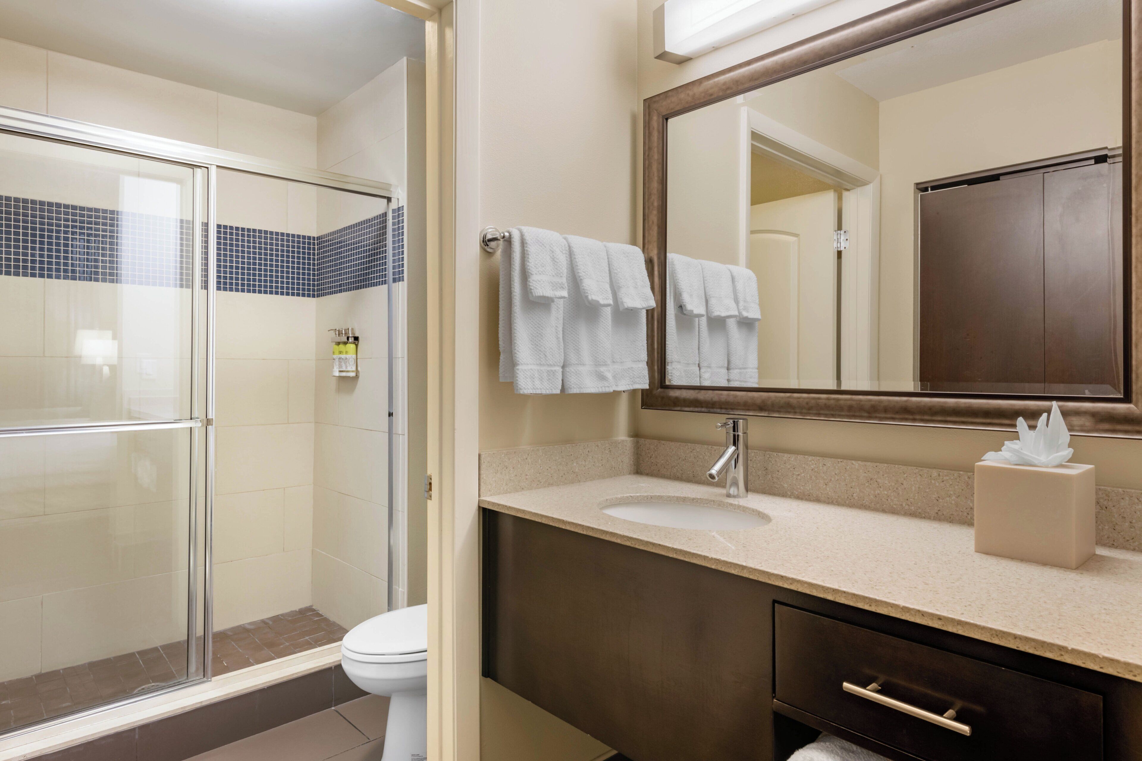 A bathroom with a glass shower enclosure, a white toilet, a sink with a mirror above it, and a towel rack with towels.