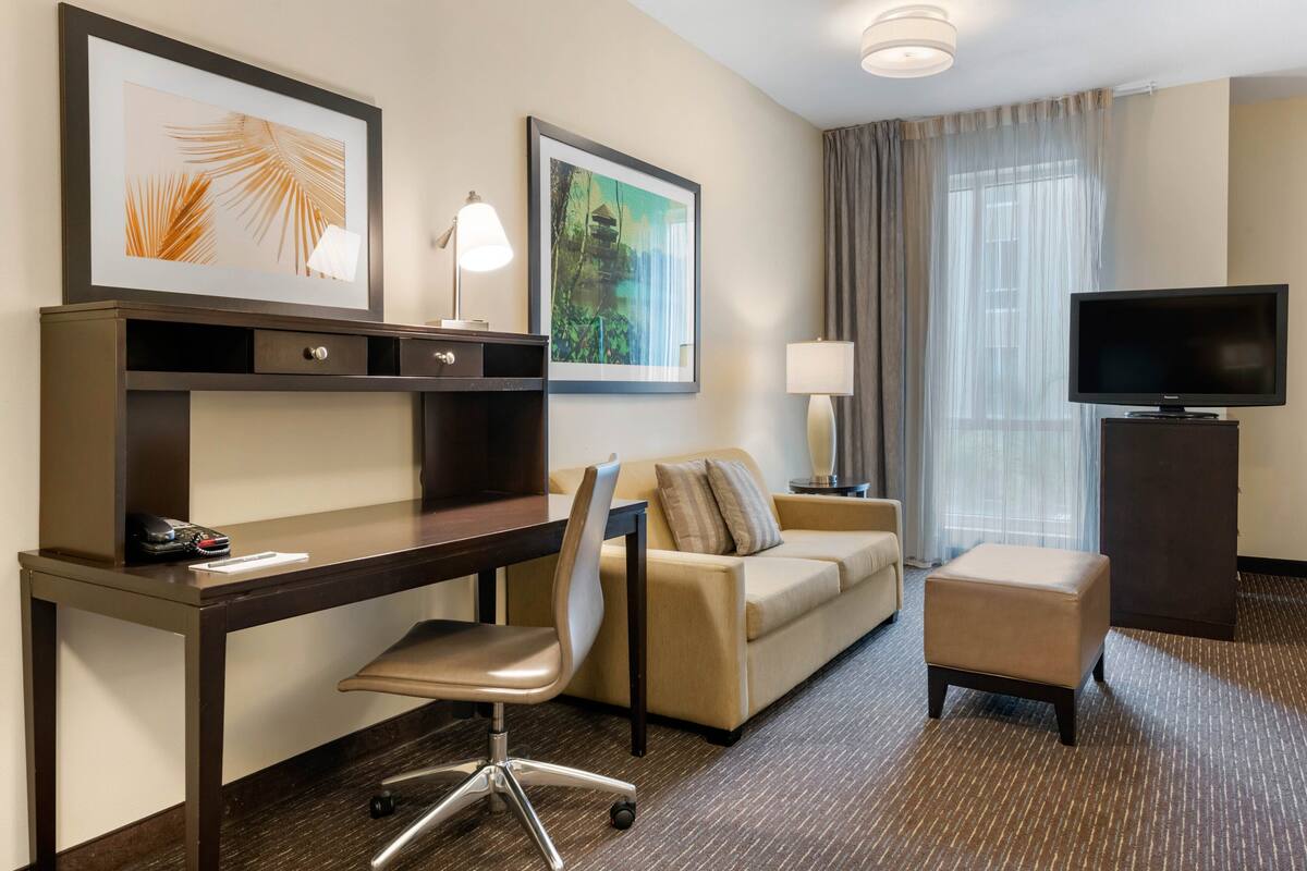 A hotel room with a desk, chair, sofa, ottoman, TV, and artwork on the walls.