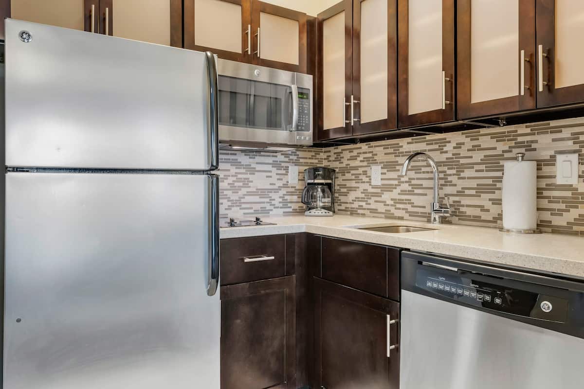 A kitchen with stainless steel appliances, dark wooden cabinets, a tiled backsplash, and a dishwasher.