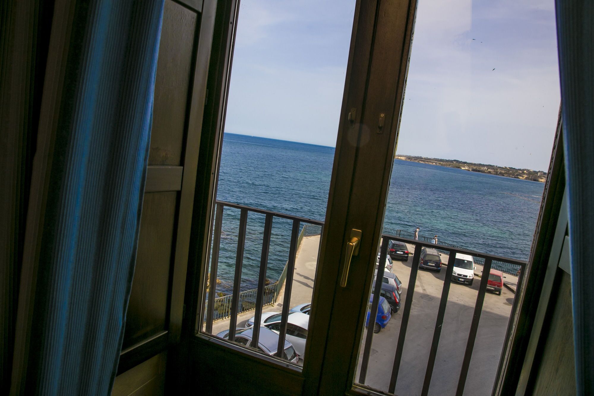 Blick vom Balkon auf ein Gewässer mit parkenden Autos und einer fernen Küste.