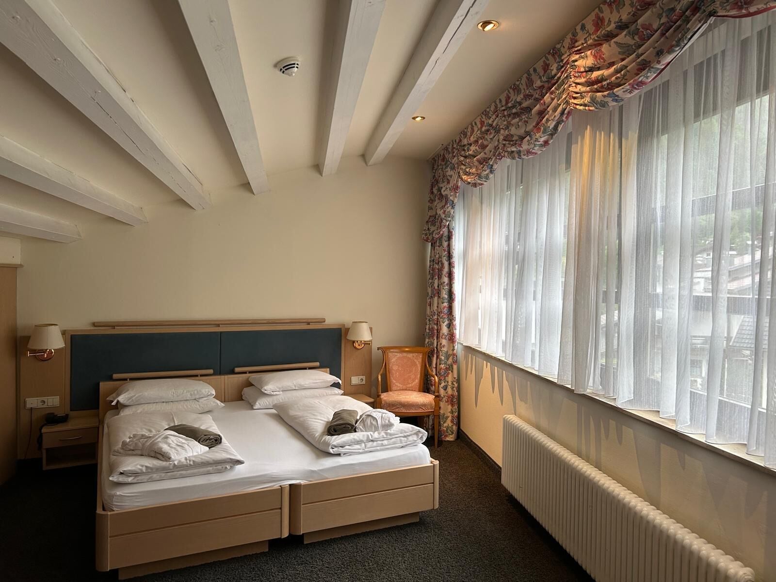 Ein Hotelzimmer mit Doppelbett, einem Sessel, einem Schreibtisch, einer Lampe und einem Fenster mit Vorhängen.