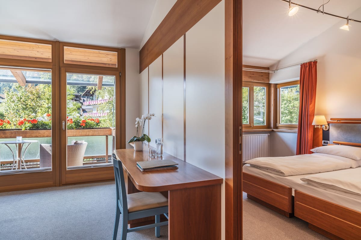A bedroom with a wooden desk, a bed, and a view of a garden through large windows.