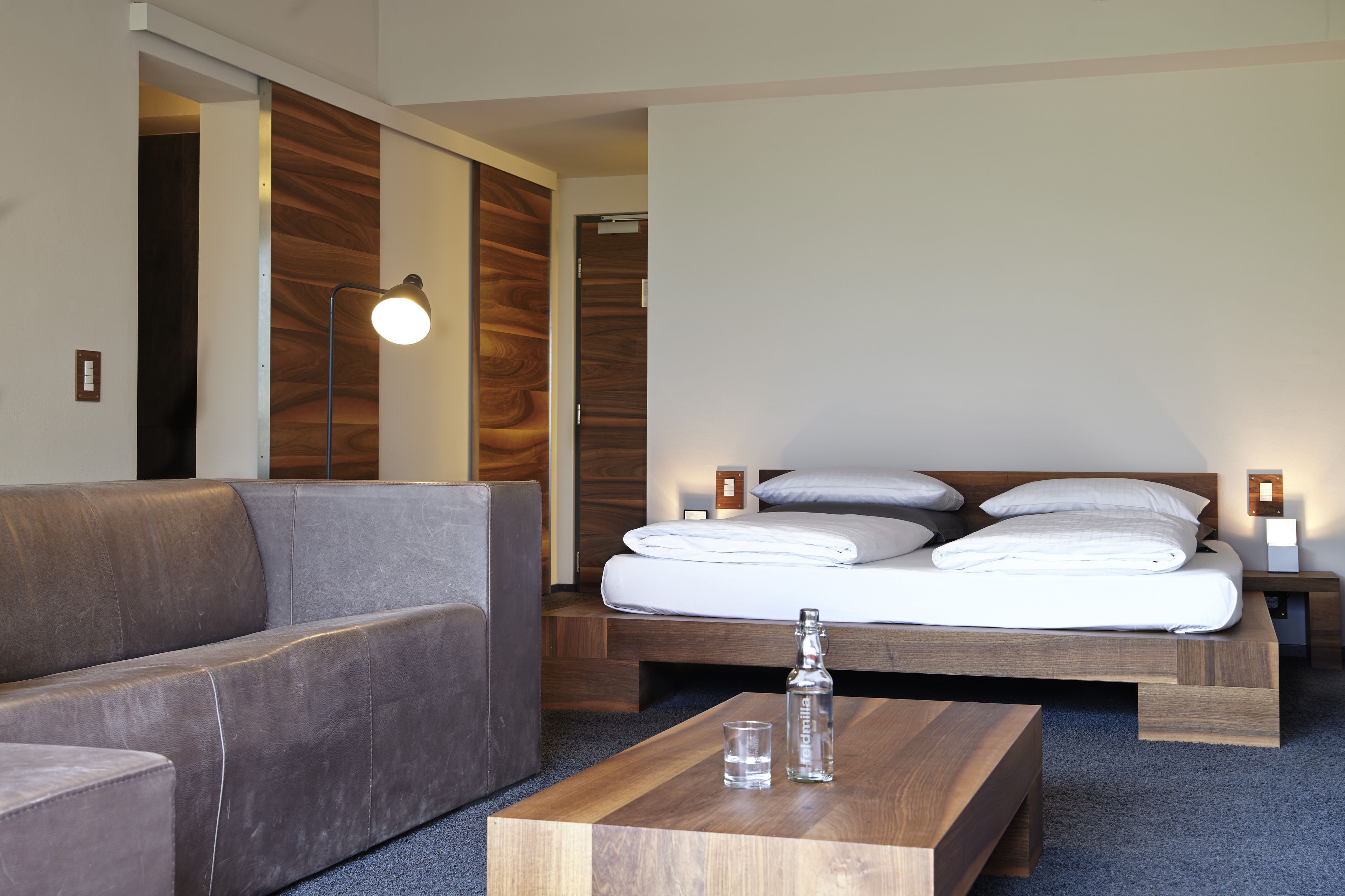 A modern hotel room with a sofa, a wooden bed, a bedside table with a bottle and glass, and a wall-mounted lamp.