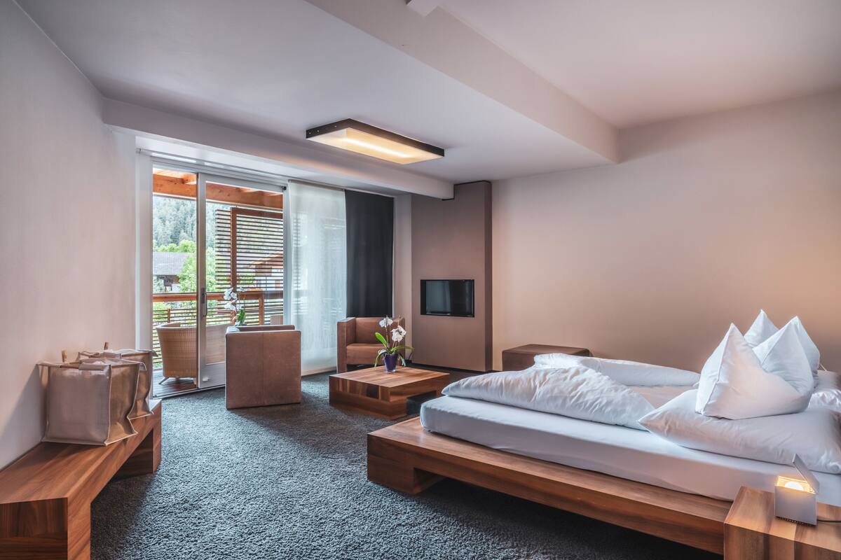 A modern hotel room with a large bed, a sofa, a coffee table, and a TV. There is a sliding glass door leading to an outdoor area.