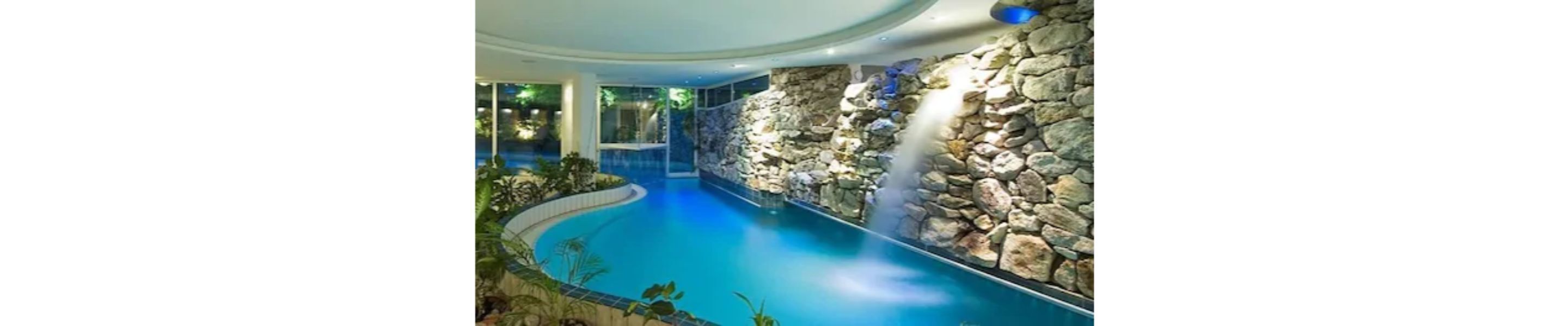 An indoor swimming pool with a curved design, surrounded by a stone wall and tropical plants.