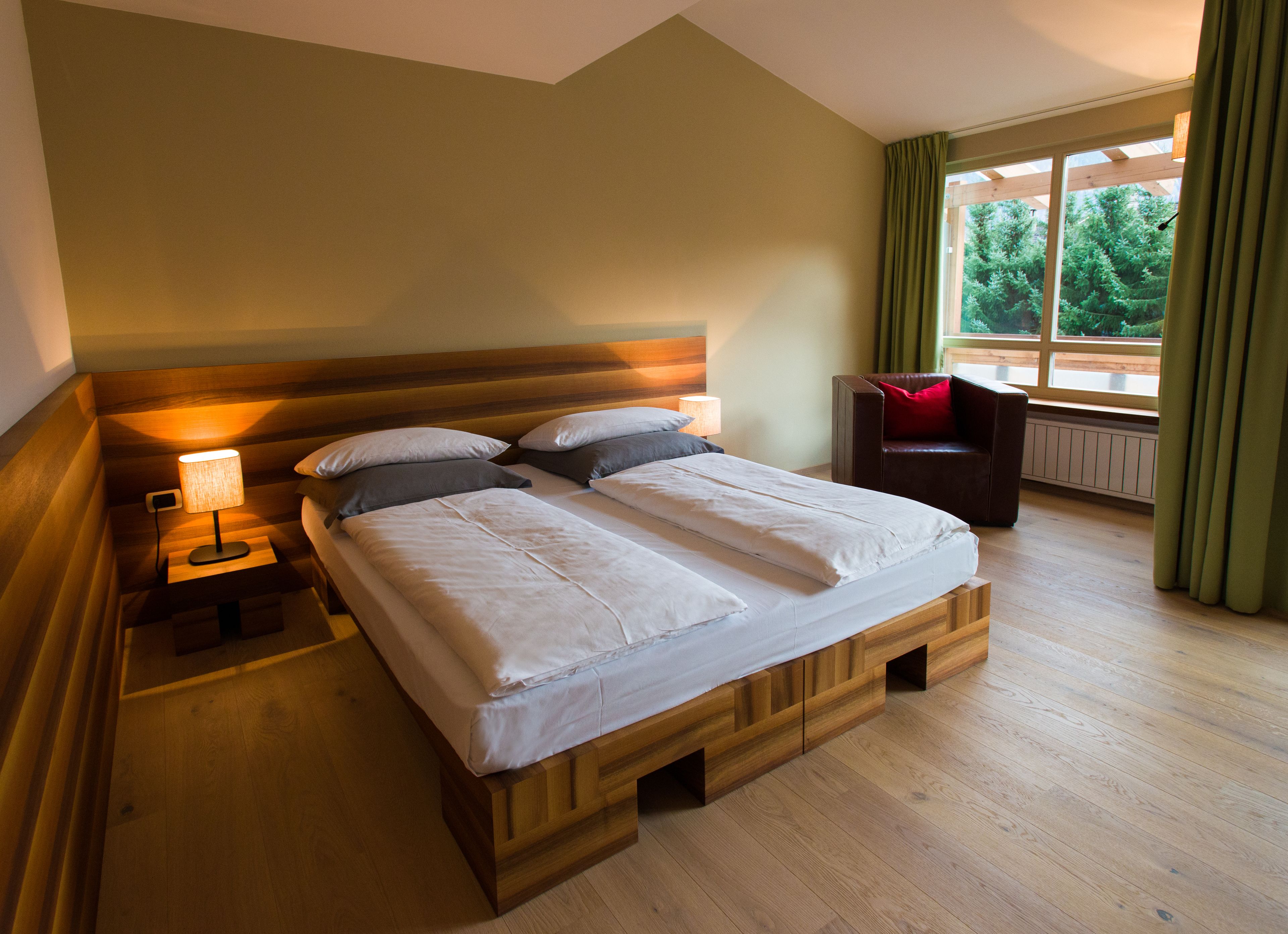 A double bed with a wooden frame, a bedside table with a lamp, and a view of greenery through a window.