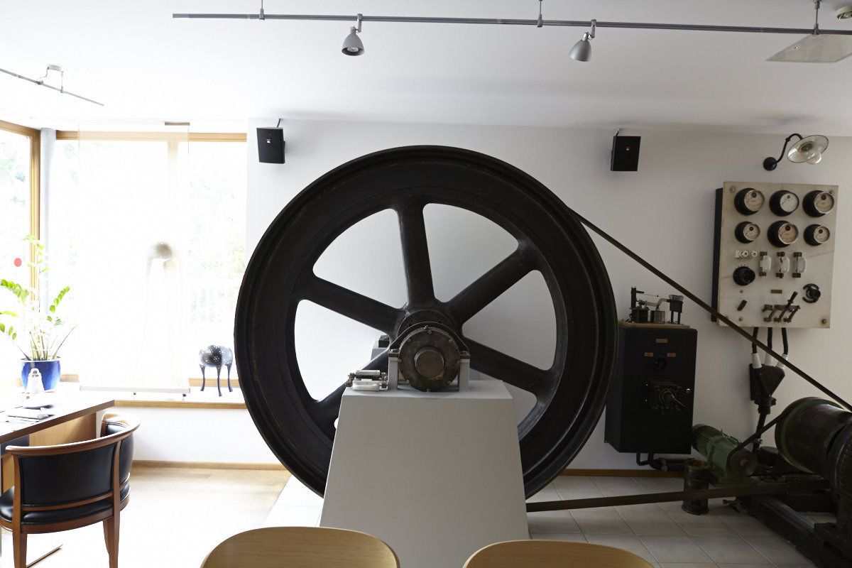 A room with a large mechanical wheel, a table with chairs, a potted plant, and a window with natural light.