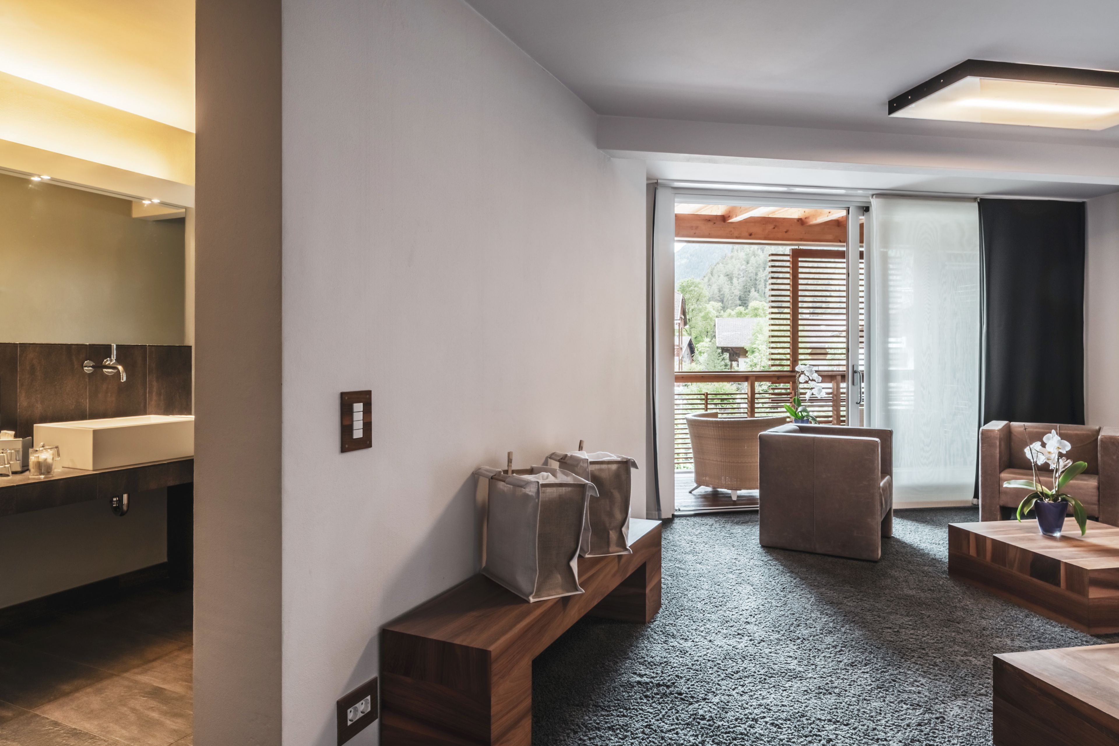 Une chambre d’hôtel moderne avec une moquette sombre, des meubles en bois, une grande fenêtre donnant sur un paysage verdoyant et une salle de bain bien éclairée.