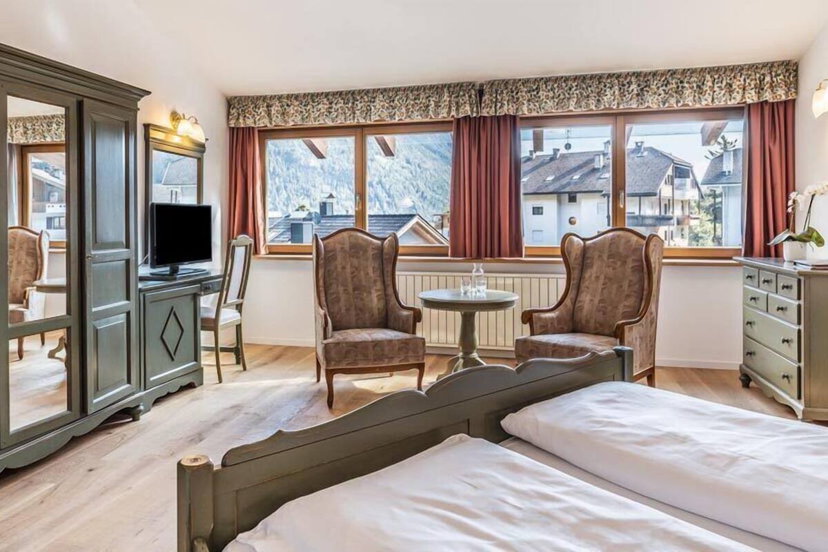 A hotel room with two beds, a desk with a computer, two armchairs, a dresser, and a window with a view of buildings.