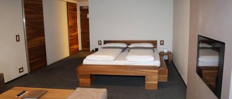 A hotel room with a wooden bed, a small bedside table, a bedside lamp, a wooden bench, and a wall-mounted TV.
