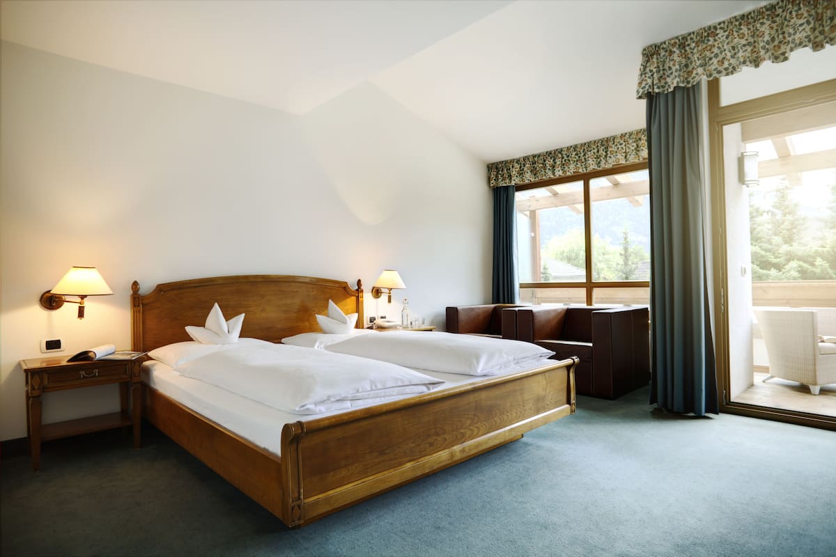 A hotel room with a wooden bed, two bedside lamps, a nightstand, a window with curtains, and a view of greenery outside.
