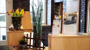 Un mostrador de recepción con acabado de madera, una planta en maceta, un florero alto con follaje verde y folletos informativos.