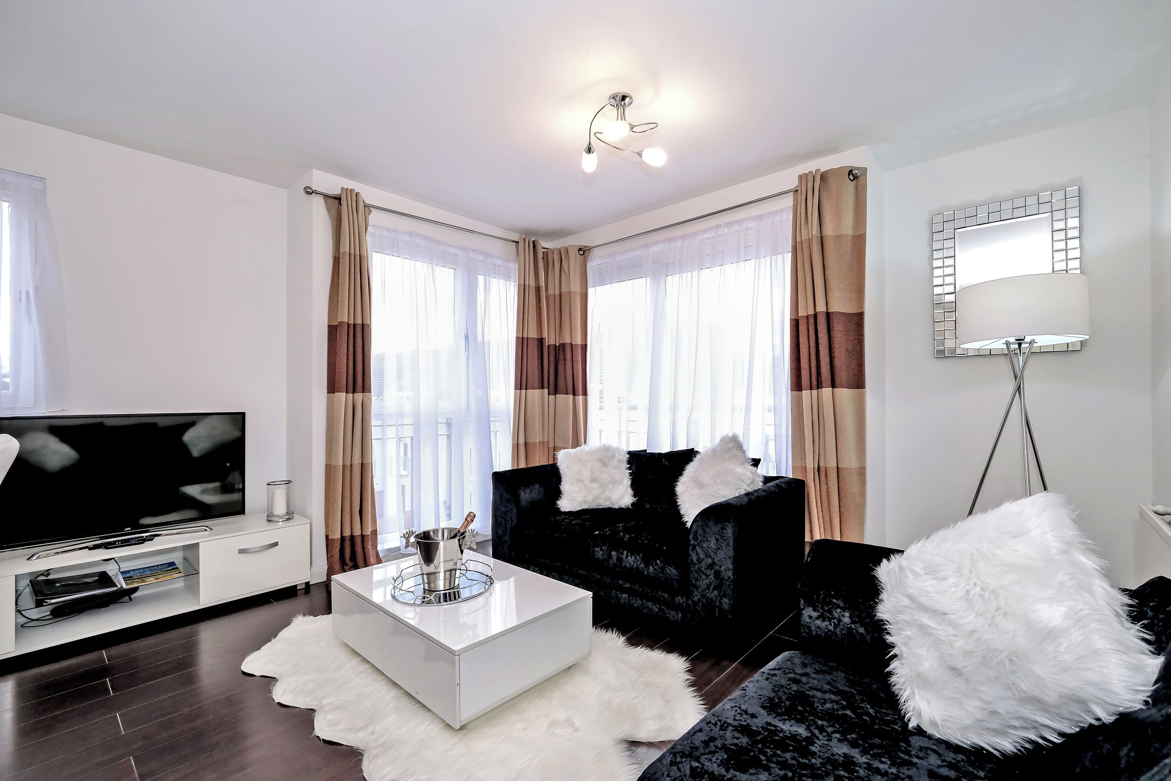 A modern living room with a flat-screen TV, a white coffee table, black sofas with white fluffy cushions, and a large mirror on the wall.