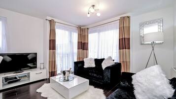 A modern living room with a flat-screen TV, a white coffee table, black sofas with white fluffy cushions, and a large mirror on the wall.