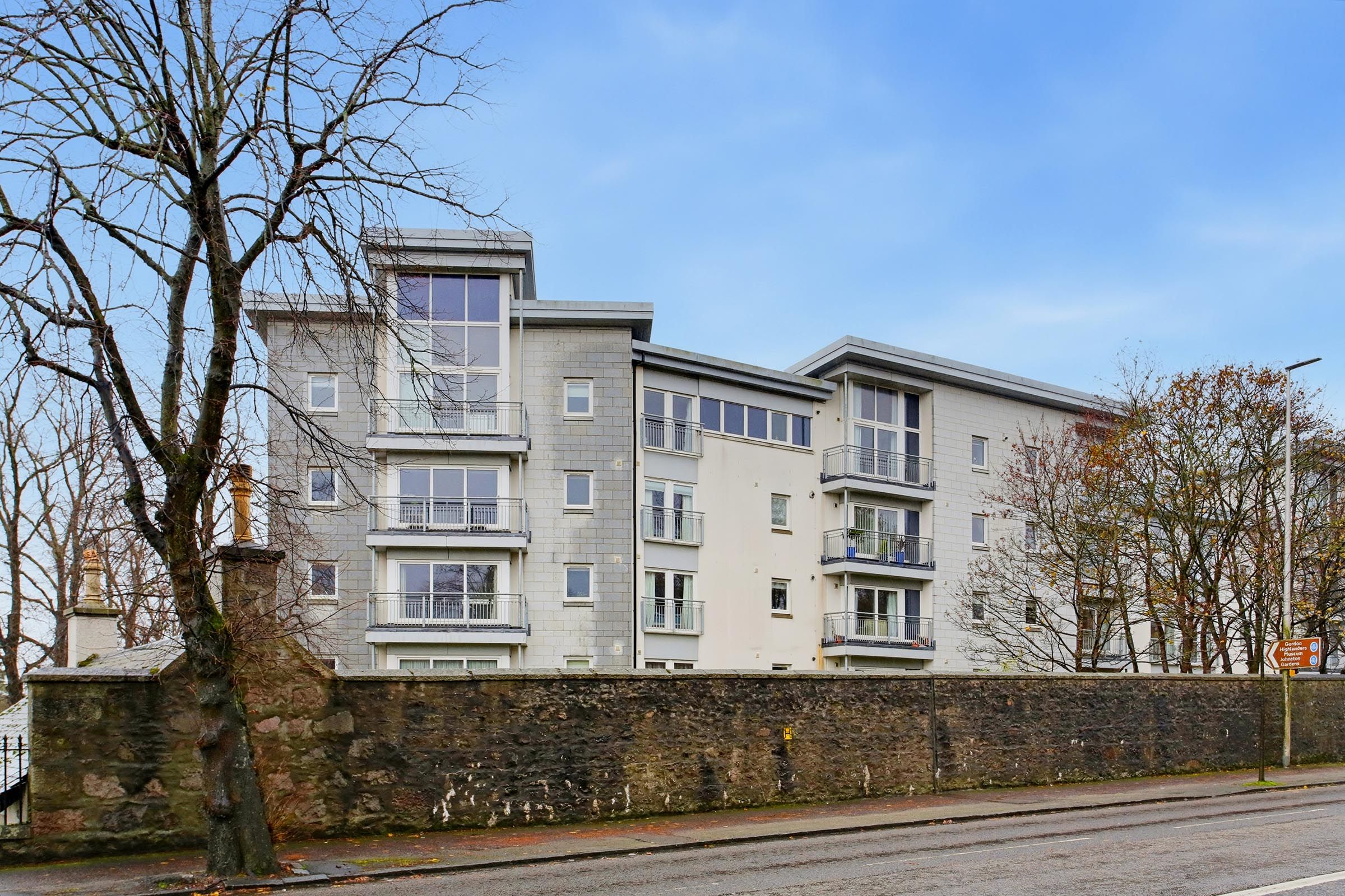 A multi-story residential building with a stone wall in front, a clear sky, and leafless trees.