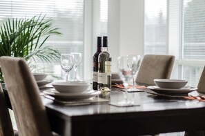 A dining table set with plates, glasses, and a bottle of wine, with a potted plant and window blinds in the background. - Orange Apartments-Kepplestone (Aberdeen)