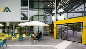 Een terras met gele en blauwe stoelen en witte parasols, voor een modern gebouw met grote ramen en een gele muur.