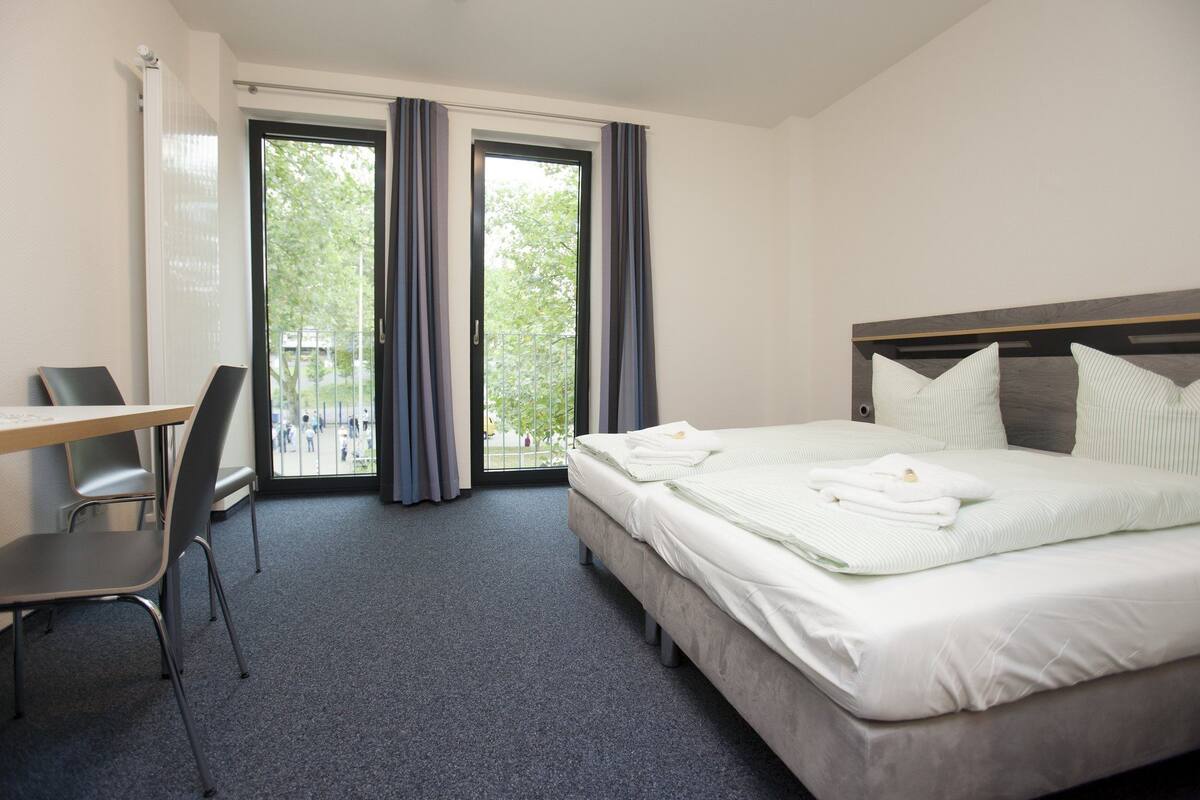 Habitación de hotel con cama, escritorio y silla. Tiene una ventana con cortinas y vistas a árboles.