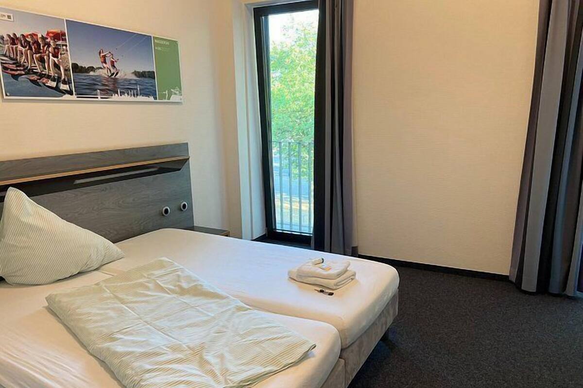 Habitación de hotel con cama, almohada, toalla doblada, ventana con cortinas y pared con cuadros.