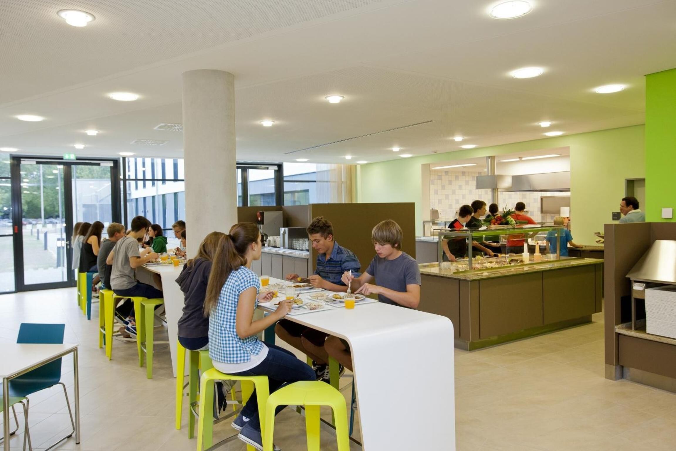 Una cafetería moderna con mesas altas y taburetes amarillo brillante, que cuenta con una zona de servicio central y amplios ventanales.