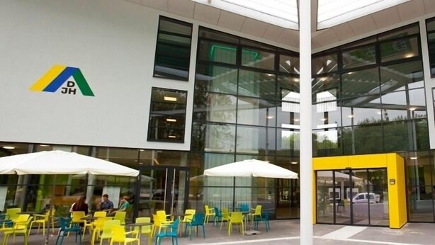 Een modern gebouw met grote glasramen, terras met gele stoelen en witte parasols, en logo's op de muren.