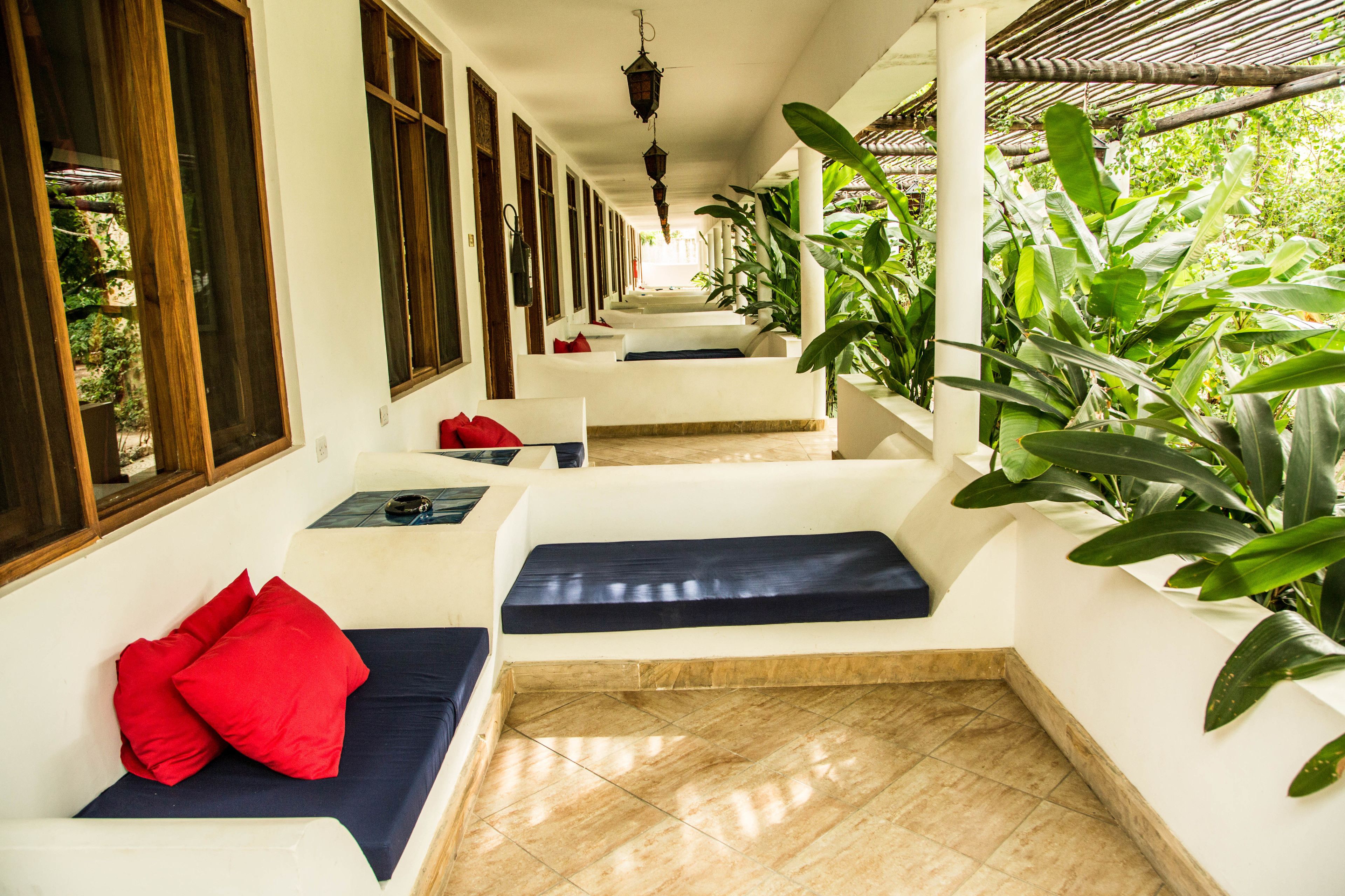 A corridor with white walls, wooden-framed windows, and a tiled floor. There are benches with cushions and a hanging lantern.