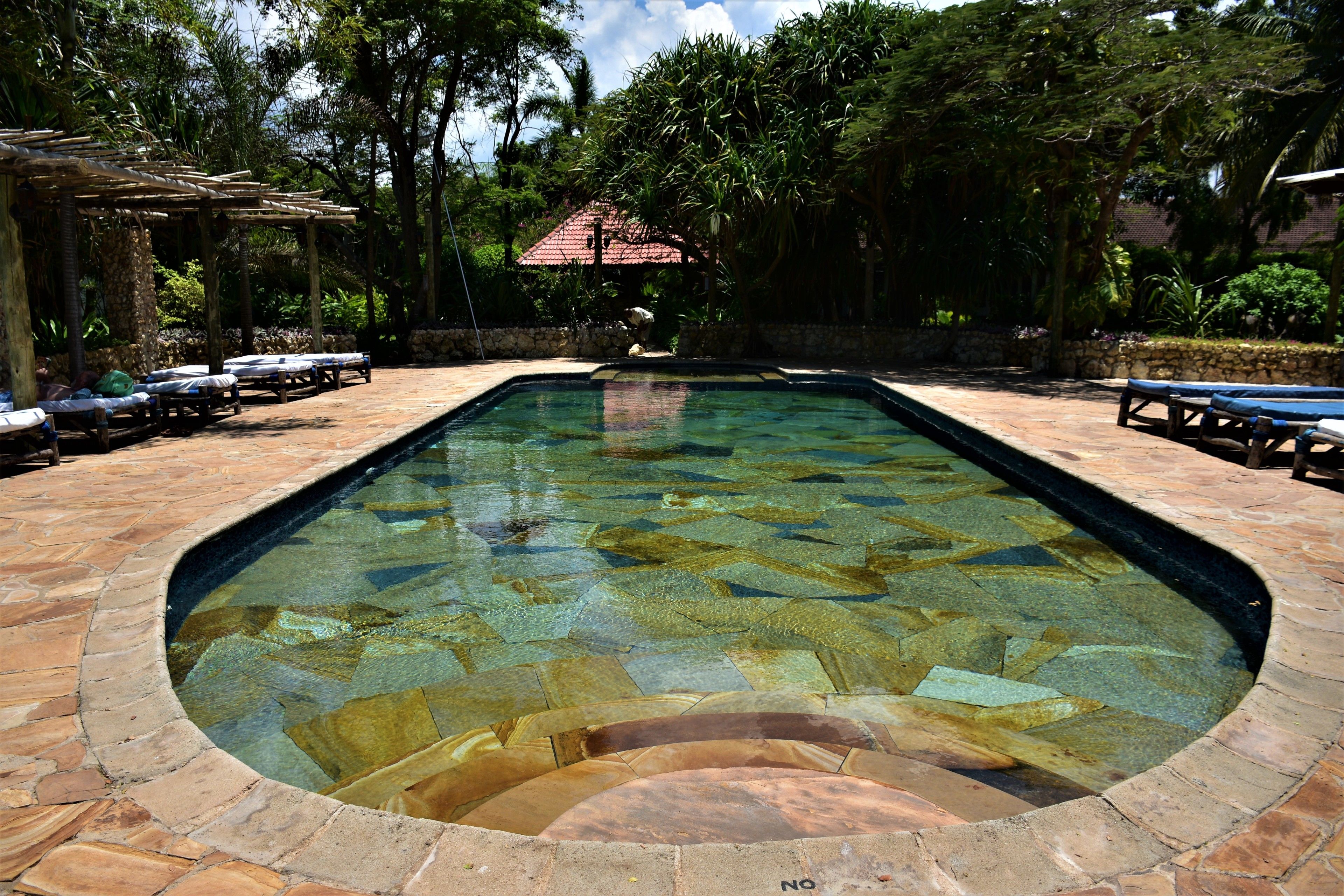 A rectangular swimming pool with clear water, surrounded by stone tiles and wooden lounge chairs.