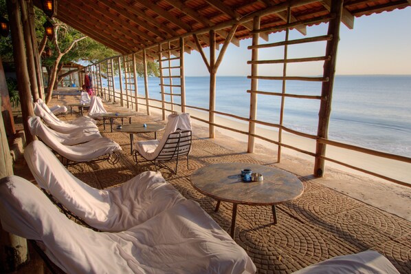 Un salón junto a la playa con asientos y mesas acolchadas de color blanco, con vistas al mar.