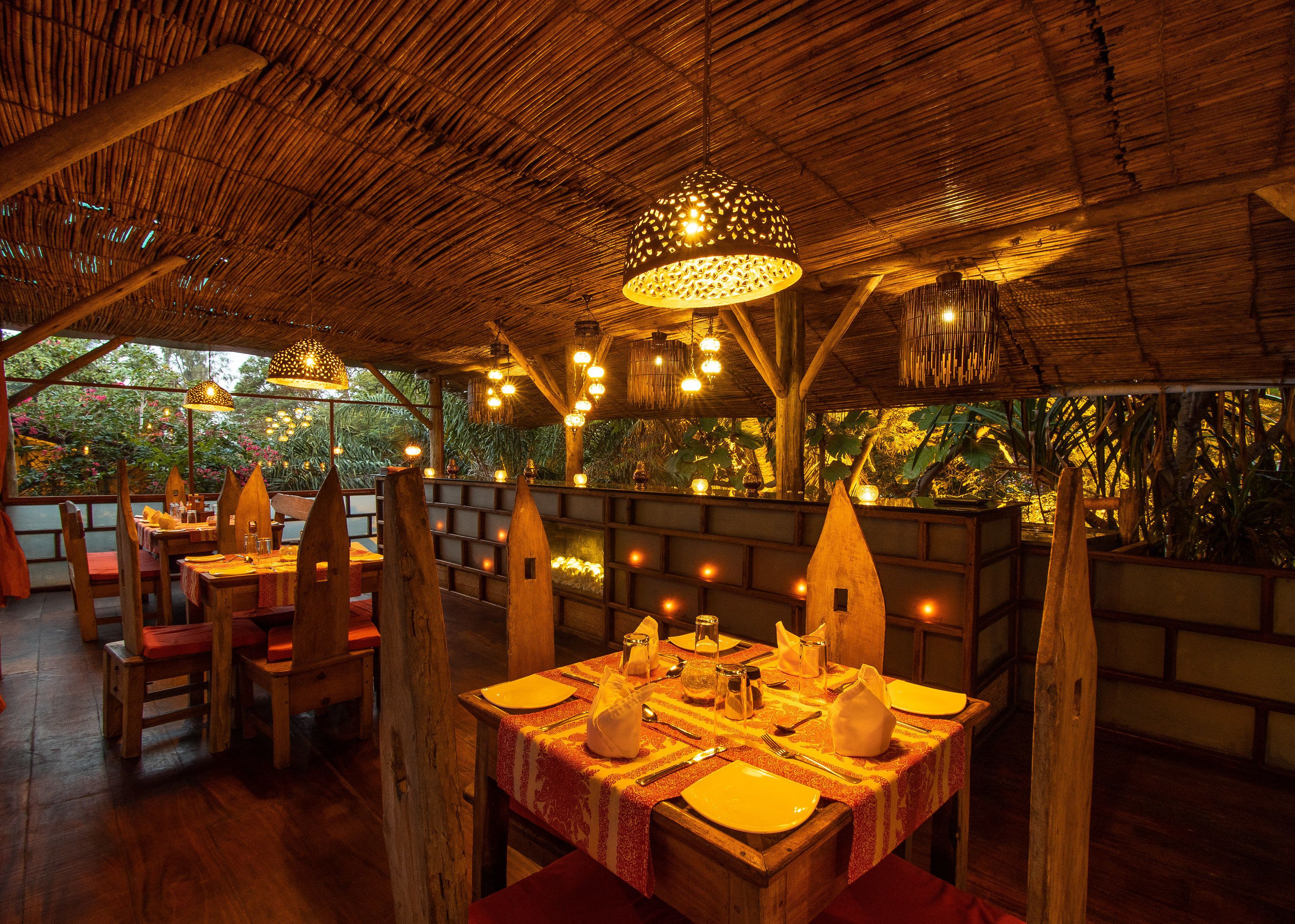 An outdoor dining area with wooden tables and chairs, surrounded by a thatched roof and illuminated by hanging lights.