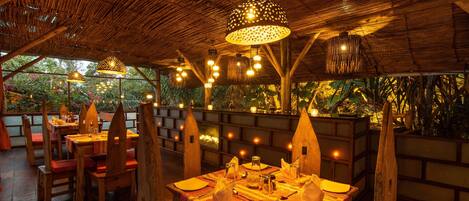An outdoor dining area with wooden tables and chairs, surrounded by a thatched roof and illuminated by hanging lights.